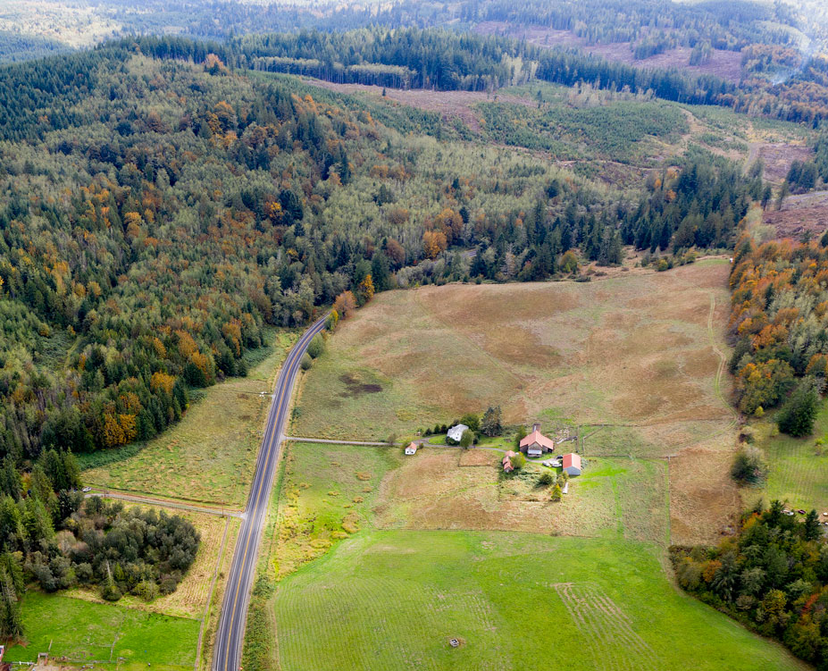 Aerial drone shot of open green farmland with some farm buildings, and forested hills beyond.