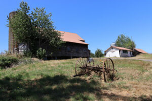 A piece of rusted farm equipment next to a weathered barn under a blue summer sky.