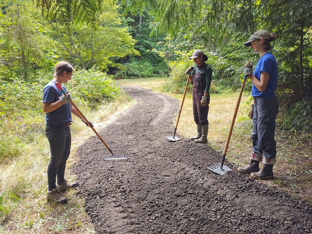 Three women with tools work on gravel trail curving through field